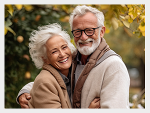 Starsza para obejmująca się i uśmiechająca do aparatu, stojąca na zewnątrz w jesiennym otoczeniu z drzewami i złotymi liśćmi w tle.